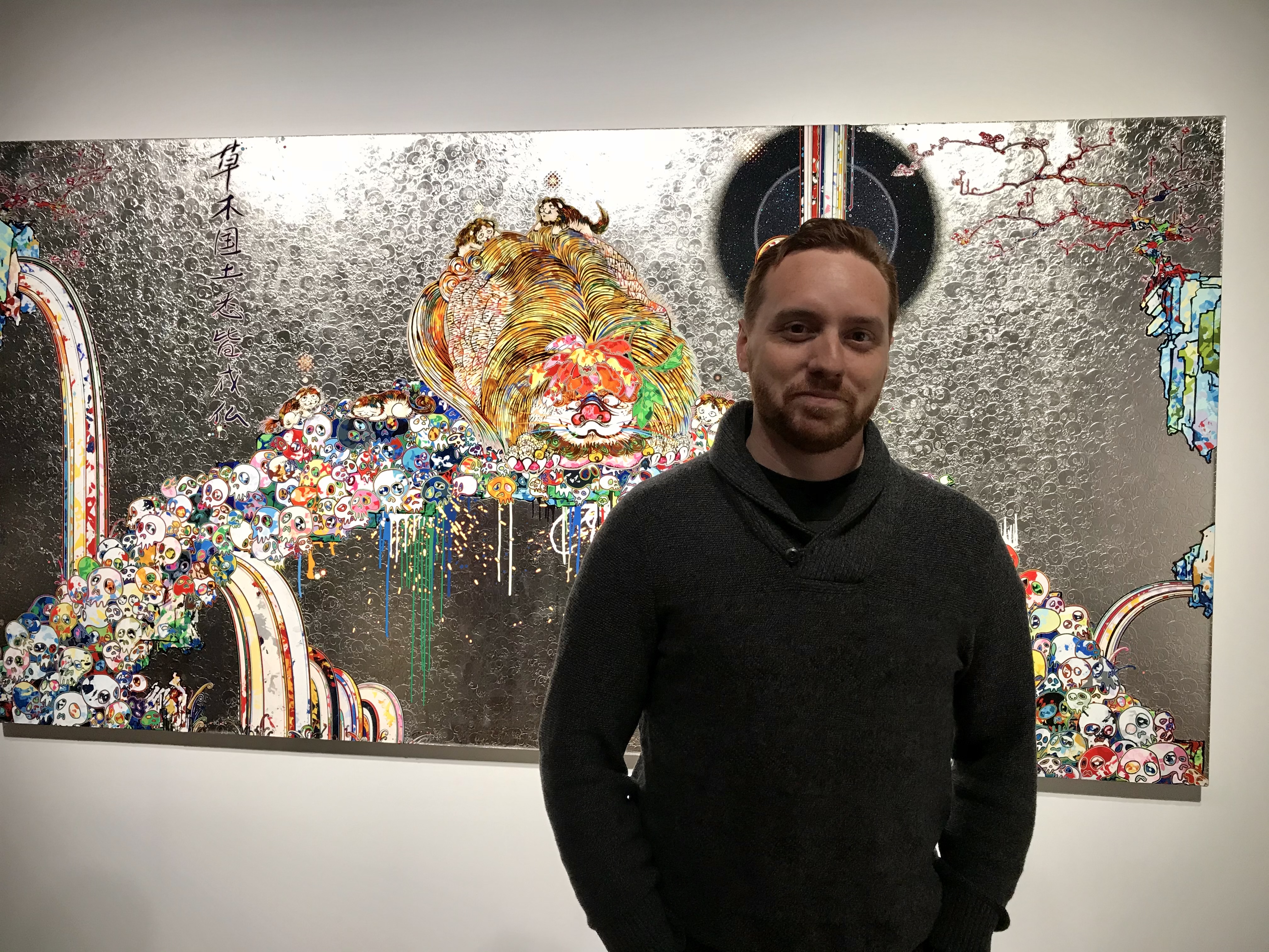 Standing in a dark sweater in front of colorful silver gallery artwork.